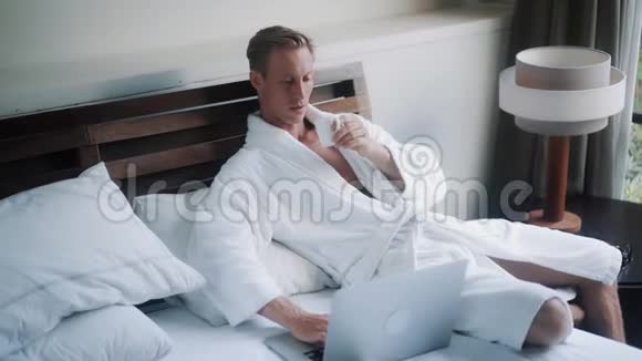 穿浴袍的年轻人在笔记本电脑附近的床上喝咖啡视频的预览图