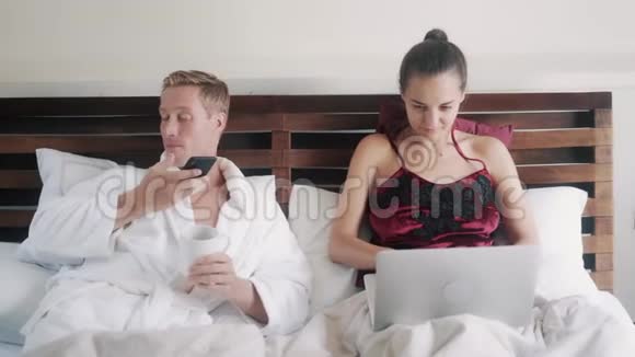 一对夫妇躺在床上男人喝咖啡用手机女人在笔记本电脑上工作视频的预览图
