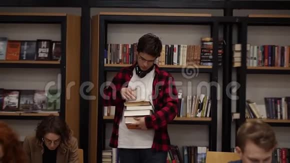 一个穿着格子衬衫和耳机的人拿着一堆书和读物一排排地走着的慢镜头其他学生视频的预览图