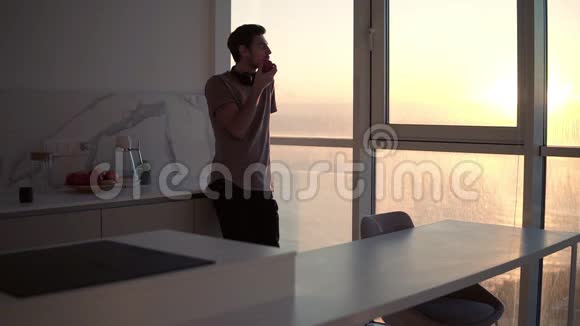 高个子年轻的单身男子带着耳机脖子上戴着耳机在工作室厨房的窗户旁放松看日落吃东西视频的预览图