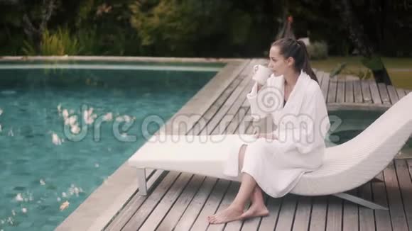 穿着白色浴袍的女人坐在靠近游泳池的日光浴床上清晨喝咖啡视频的预览图