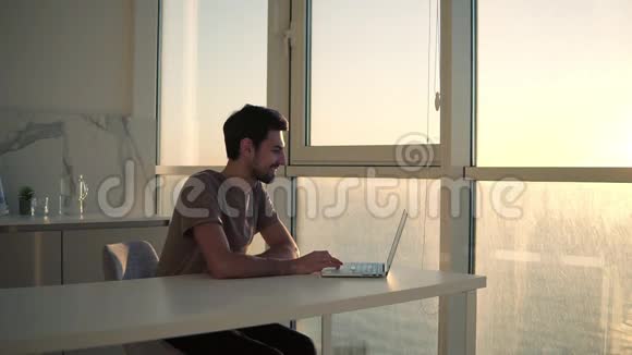 欧洲深色头发的男人坐在工作室厨房的全景窗口附近看着笔记本电脑桌面兴高采烈视频的预览图