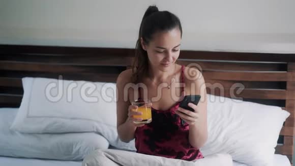 美丽的女孩带着一杯果汁冲浪网在大床上有智能手机视频的预览图