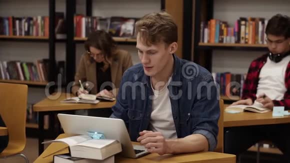 金发白种人坐在大学图书馆里用笔记本电脑工作准备考试然后看着相机视频的预览图
