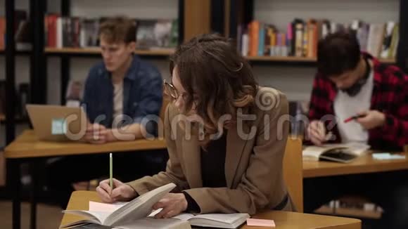 一个穿着棕色夹克的古丽的欧洲女孩坐在大学图书馆里写论文准备考试然后看上去视频的预览图