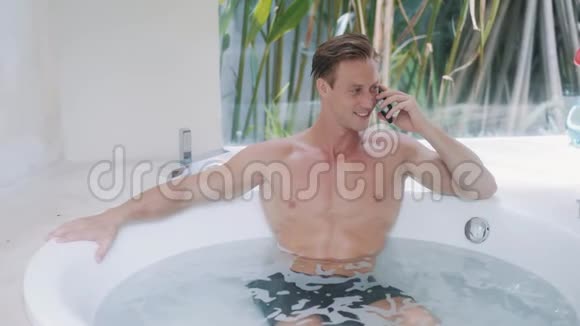 快乐男人坐在家里的浴缸里谈论智能手机视频的预览图