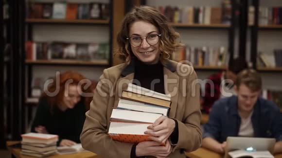 吸引人的欧洲女学生拿着书在高中图书馆微笑着看相机的肖像教育视频的预览图