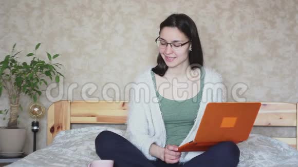 年轻迷人的女孩正在喝一杯粉红色的咖啡而她的笔记本电脑坐在家里的床上视频的预览图