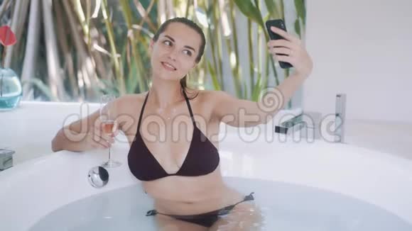 穿比基尼带香槟的快乐女人在浴缸里自拍视频的预览图