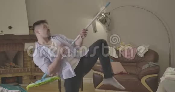 开朗的白种人模仿在拖把上弹吉他快乐的帅哥在家里打扫卫生时玩得很开心视频的预览图