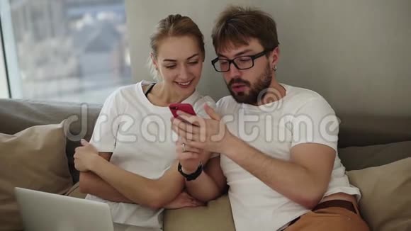 漂亮的年轻夫妇坐在客厅的沙发上坐在现代化的阁楼内部一起看智能手机视频的预览图