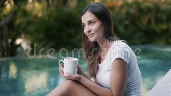 漂亮的黑发女人从游泳池的杯子里喝茶视频的预览图