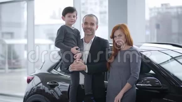 快乐的白人家庭站在汽车经销商看着相机和微笑年轻的父亲母亲和儿子视频的预览图