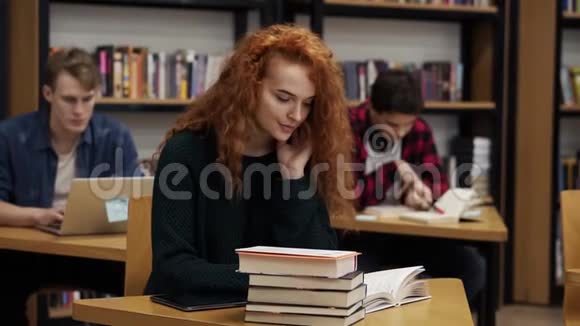 一位年轻的红头卷发女学生正在写论文或准备考试的肖像坐着看书视频的预览图
