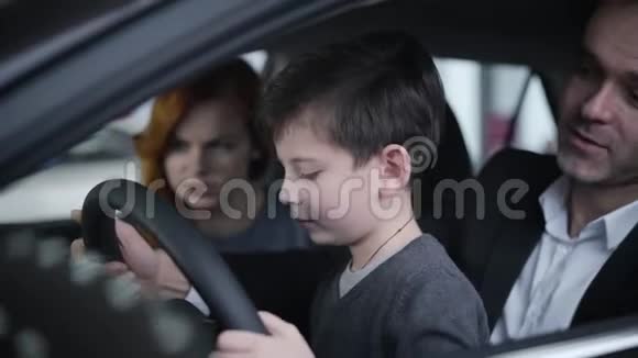 可爱的小白种人男孩和父母坐在车里拿着方向盘的特写肖像快乐成功视频的预览图