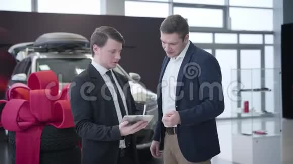 两个白种人站在汽车经销商和谈话汽车经销商持有平板电脑和咨询客户陈列室视频的预览图