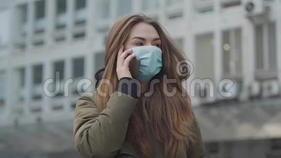 年轻漂亮的女人戴着保护面具站在城市里打电话深色头发的女孩咳嗽全球视频的预览图