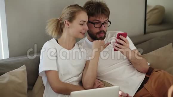 放松的年轻夫妇一起坐在客厅的沙发上看智能手机在现代阁楼内部视频的预览图