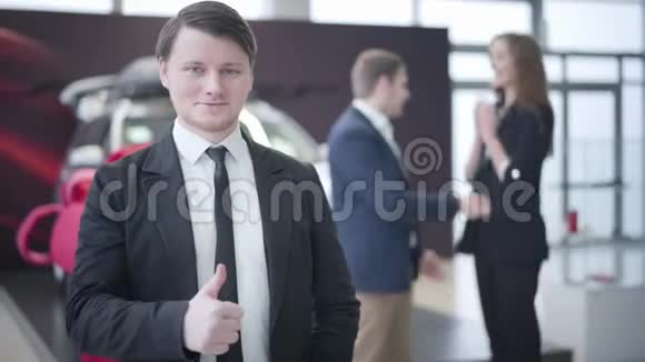 一幅自信的白种人汽车经销商站在经销商的肖像并显示拇指作为年轻的幸福夫妇拥抱和拥抱视频的预览图