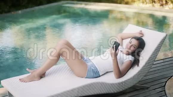 女人躺在靠近游泳池的日光浴床上用电话放松享受假期视频的预览图