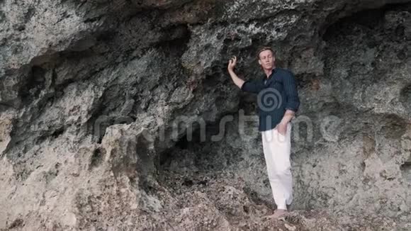 穿着白色裤子和黑色衬衫的白人男子站在靠近岩石的海滩上向远处望去视频的预览图