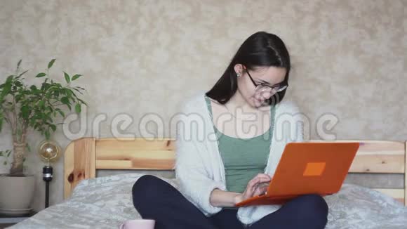 年轻迷人的女孩正积极浏览笔记本电脑上的新闻而坐在家里的床上视频的预览图