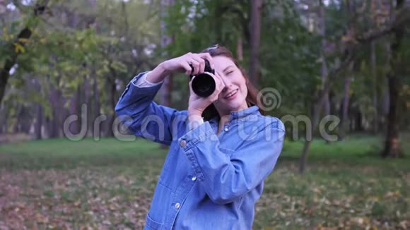 可爱的时髦摄影师女孩在专业相机上拍照并微笑秋天公园的肖像反对视频的预览图