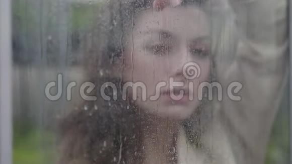 布鲁内特白种人女人站在窗户后面看着摄像机漂亮的女士独自度过雨天照相机视频的预览图
