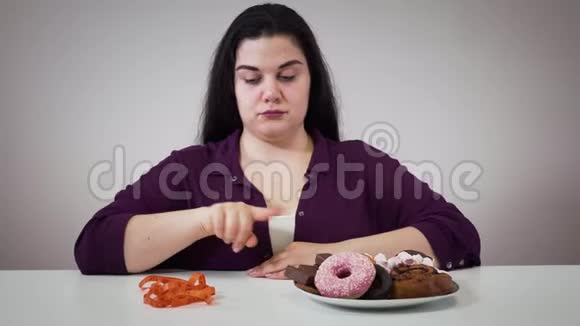 丰满悲伤的高加索女人坐在糖果和测量磁带前的肖像胖女孩在旁边移动盘子视频的预览图