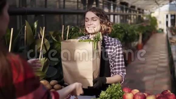 欧洲女售货员穿着围裙的肖像正在用棕色纸袋将有机食品送给温室里的女顾客视频的预览图