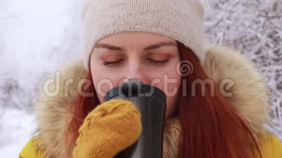 年轻的红发女孩戴着一顶温暖的针织帽子和一件黄色的冬季夹克在金属杯上喝一杯热饮料视频的预览图