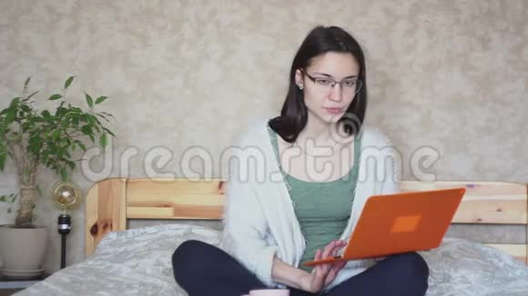 年轻迷人的女孩正在喝一杯粉红色的咖啡而她的笔记本电脑坐在家里的床上视频的预览图