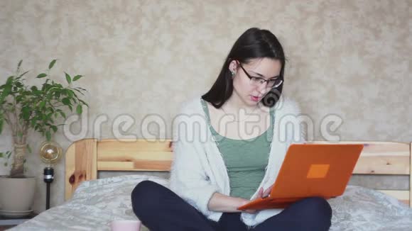 年轻迷人的女孩正积极浏览笔记本电脑上的新闻而坐在家里的床上视频的预览图