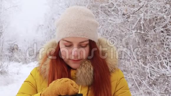 年轻的红发女孩戴着一顶温暖的针织帽子和一件黄色的冬季夹克在金属杯上喝一杯热饮料视频的预览图