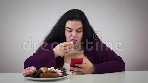 使用智能手机和吃糖果的浓白种人肥胖女孩的肖像肥胖的年轻女性超重视频的预览图
