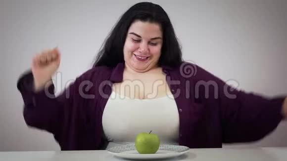 快乐肥胖白人妇女坐在桌子旁吃苹果的肖像微笑的黑发女孩吃健康低视频的预览图