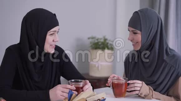两个快乐的穆斯林妇女在室内谈笑风生坐在餐桌前喝茶的女性朋友视频的预览图