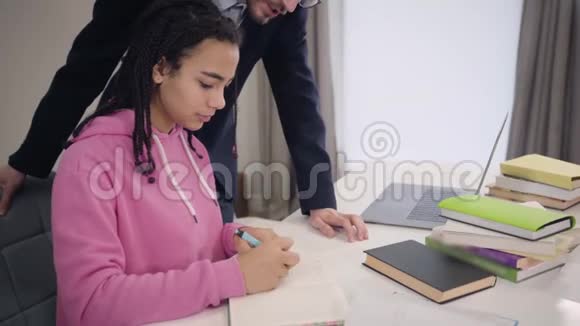 穿着悲伤的非裔美国女孩坐在桌子旁她的白人家庭教师站在桌子旁说话视频的预览图