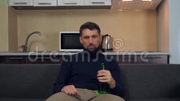 穿着深蓝色套头衫和灰色裤子的严肃的年轻人坐在沙发上喝着啤酒看电视上的娱乐节目视频的预览图