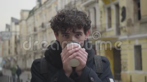 特写肖像正面英俊的白种人男孩喝咖啡在镜头前眨眼站着微笑的年轻人视频的预览图