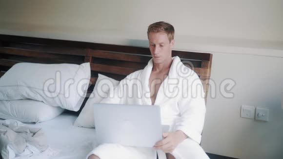 穿着白色浴袍的帅哥躺在公寓的床上用笔记本电脑上班视频的预览图