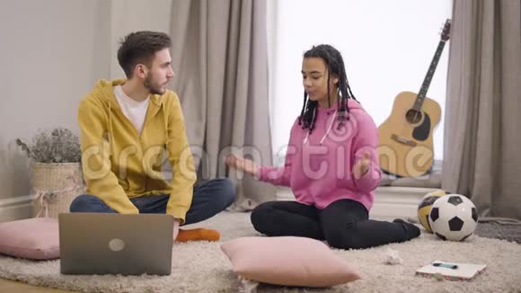 年轻的非裔美国女孩和白人男孩坐在地板上的软地毯上聊天多民族夫妇视频的预览图