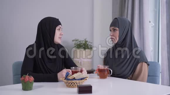 两个穆斯林妇女戴着头巾端着茶坐在桌旁说话积极的年轻女士在室内闲聊东部视频的预览图