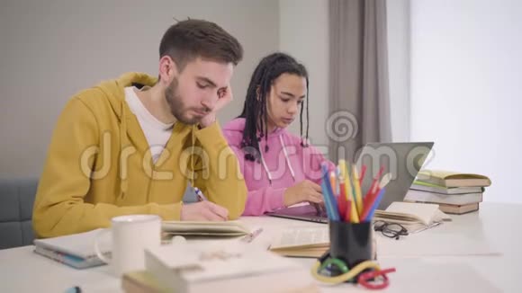 镜头接近坐在桌子旁学习的年轻多民族学生的脸无聊的白人男孩和视频的预览图