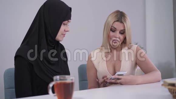 自信的年轻高加索女人向穆斯林朋友展示智能手机的肖像戴头巾的保守女人视频的预览图