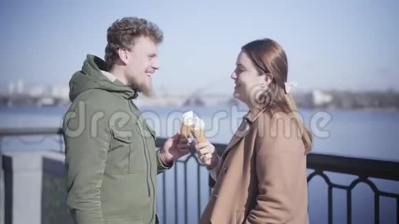 积极的白种人男朋友和女朋友拿着冰淇淋在户外聊天快乐的年轻夫妇在河边约会视频的预览图