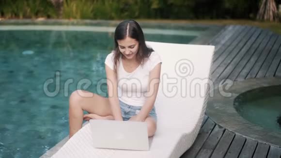 快乐的女孩坐在游泳池旁的沙发上工作视频的预览图