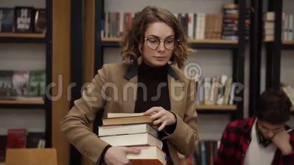 一个穿着棕色夹克和眼镜的女孩拿着一堆书从书架上走过来的慢镜头视频的预览图