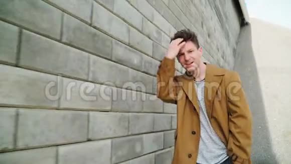 时尚美丽的年轻快乐的白种人沿着城墙微笑他穿着一件布朗朗外套关门视频的预览图