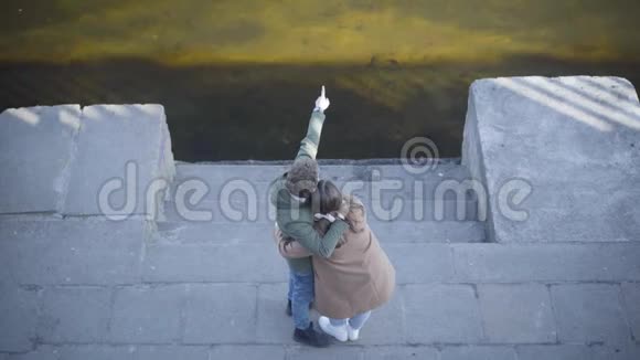 站在河岸边聊天的年轻情侣的绝景拥抱男人和女人在阳光下户外聊天视频的预览图